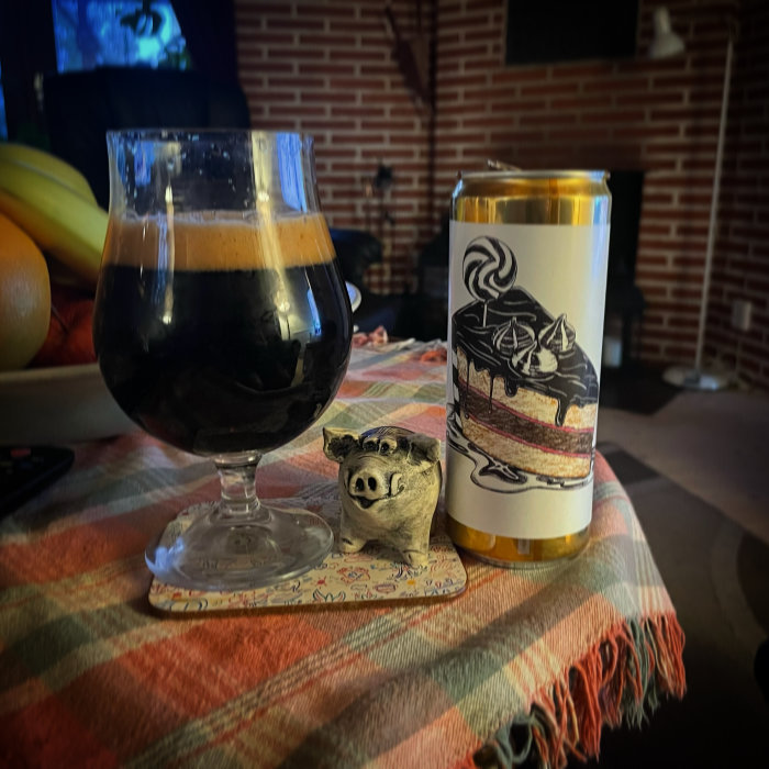 Mörk öl i ett glas på ett mönstrat underlägg bredvid en burk med illustration, framför en tänd kamin.