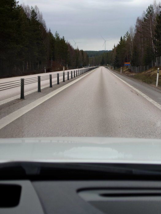 Vy från bilens förarsida som visar en tom landsväg med vägräcken och vindkraftverk i bakgrunden.