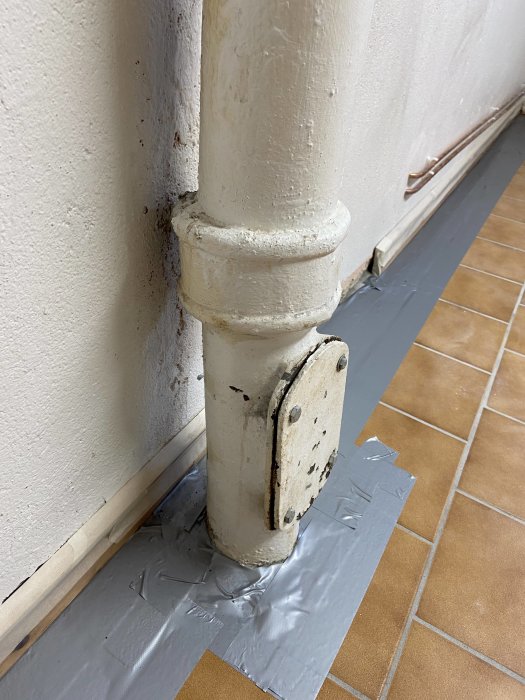 Målat rör som integreras med väggen, monterat på golv med klinker, skyddas av silvertejp vid renovering.