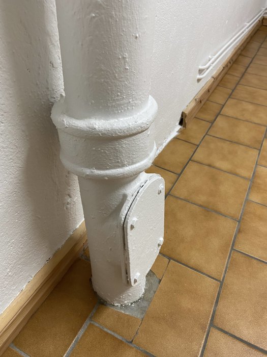 Vitmålat rör som går ner i golv med en matchande vit konsol, mot en vita väggen och terrakottafärgat golv.