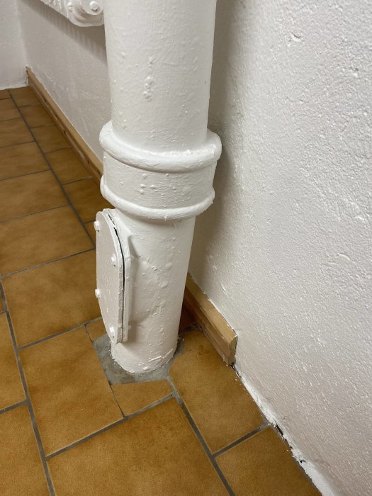 Målat vitt rör som går längs med en vit vägg och ner i ett brunt kakelgolv.