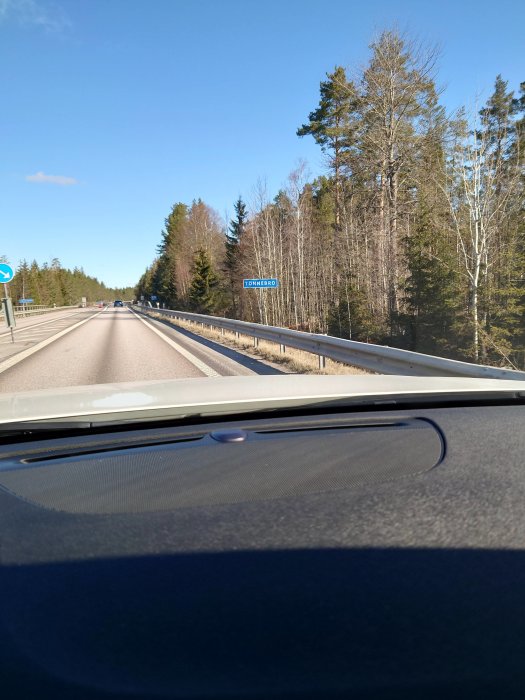 Vy från bilens förarplats på en tvåfältsväg med vägskylt och skog i bakgrunden.