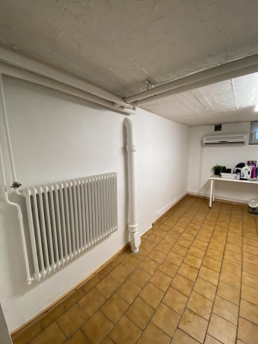 Nymålad vit radiator och rör mot en vit vägg och tak i ett rum med terrakottafärgade klinkergolv.