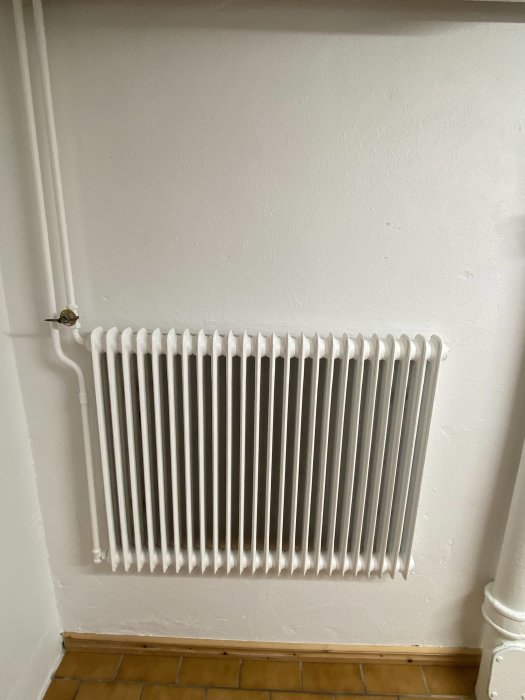 Nymålat vitt element och rör mot en vit vägg ovanför ett golv med terrakottakakel.