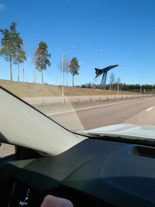 Vy från bil av vägkant med konstinstallation av upphängt flygplan på pelare mot blå himmel.