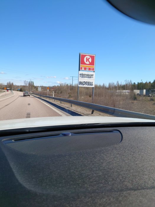 Utsikt från bilens vindruta som visar en skylt med bensinmack och hotellsymboler vid en vägkant under klar himmel.