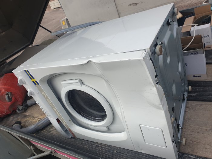 En vit tvättmaskin liggande på sidan med framsidan synlig, på en lastbilsflak.