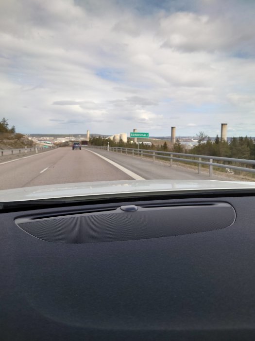 Vy från bil på motorväg mot skylt med texten "SUNDSVALL", omgiven av blå himmel och industribyggnader.