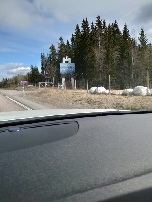 Vägskylt med texten "HÄRNÖSAND" vid sidan av en landsväg med skog i bakgrunden, sett från bilens insida.