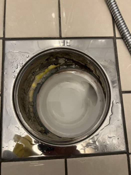 Detaljbild av en golvbrunn med misscentrerade delar och gult poröst material runt kanterna, omgiven av våta kakelplattor.