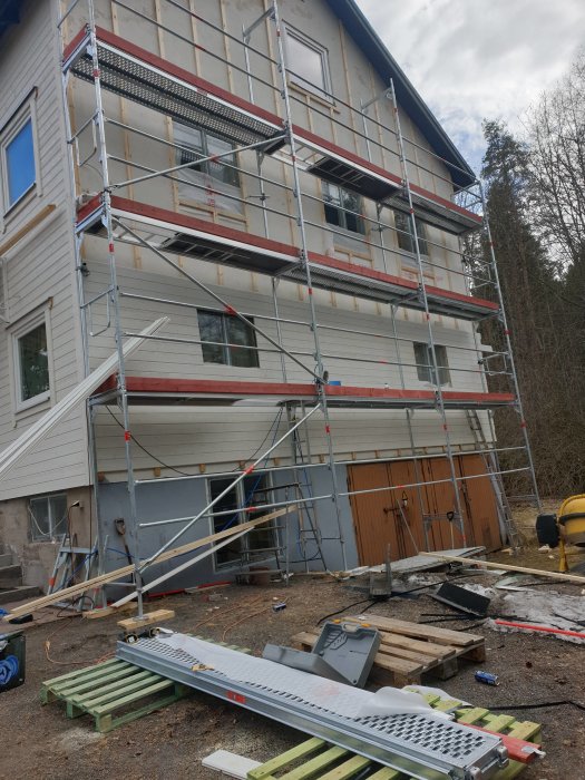 Byggarbetsplats med ställningar runt halvfärdigt isolerat hus och träpaneler på marken.