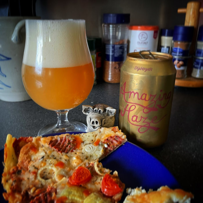 Ett glas med disig öl, en burk märkt "Amazing Haze" och en bit pizza på en blå yta med kryddburkar i bakgrunden.