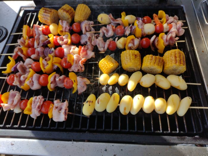 Grillade grönsaker och köttspett samt majskolvar och potatishalvor på en grill.