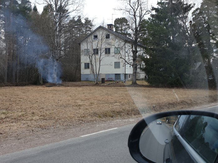 Vit hus med två våningar sedd från bil, rök syns nära huset med träd i bakgrunden och torrt gräs i förgrunden.