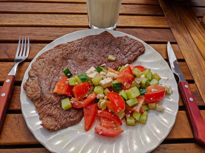 Grillad biff med tomat- och gurksallad på en tallrik bredvid ett glas mjölk på ett träbord.