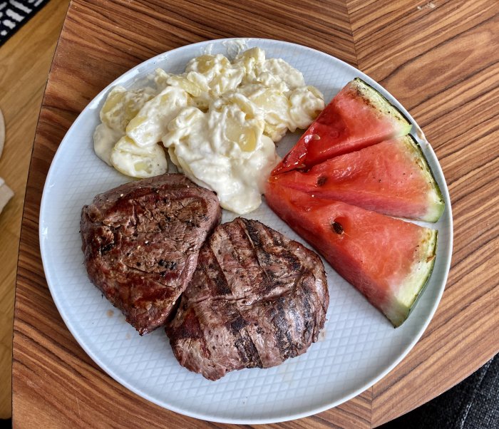 Grillade köttbitar, potatissallad och två skivor vattenmelon på en vit tallrik.