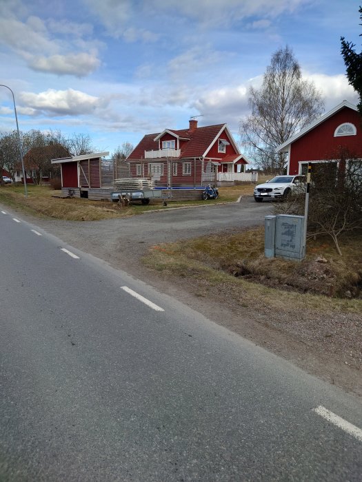 Röd trävilla i bakgrunden, med borttaget staket och vegetation före renovering, vid en asfalterad väg.