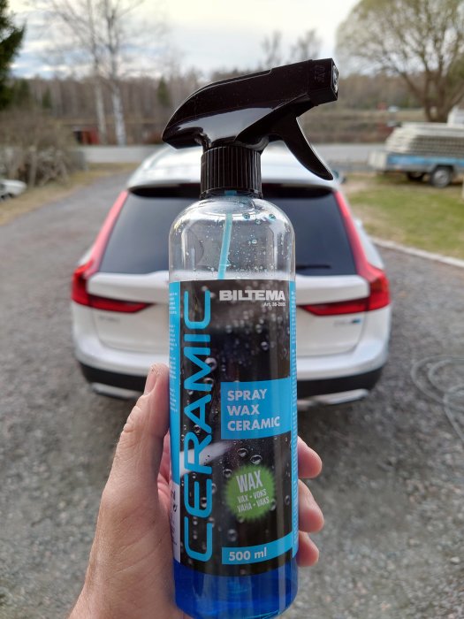 Hand håller en flaska Biltema keramisk sprayvax framför en suddig bil.
