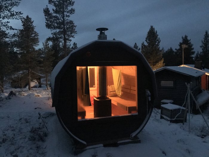 Bastutunna med tänd belysning i vinterlandskap vid skymning, skorsten i förgrunden.