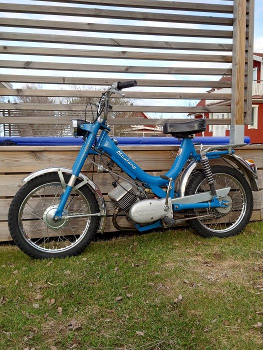 En blå moped parkerad på en gräsmatta framför en träaltan och ett rött hus i bakgrunden.