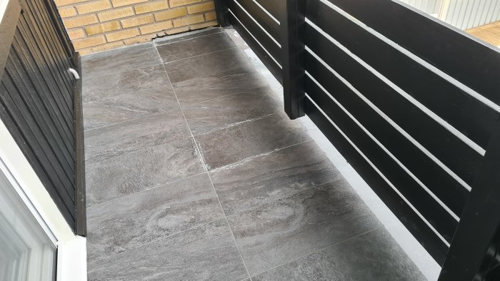 Balkonggolv med mörkgrå klinkerplattor och en svart staketstolpe, detaljer av tätning nära en vit byggnadsvägg.