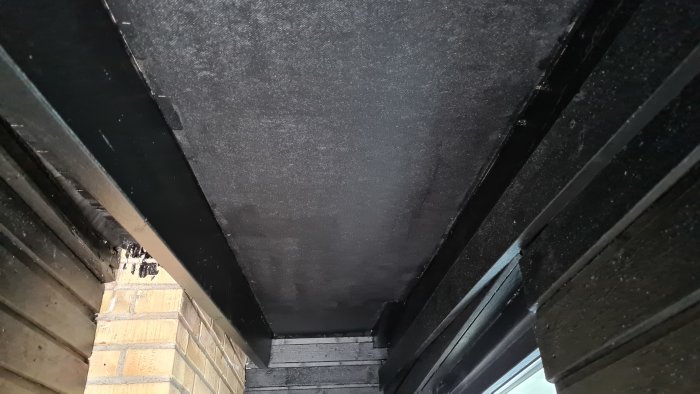 Utsikt under balkong med svart målad masonit och synliga balkar samt takkanter.