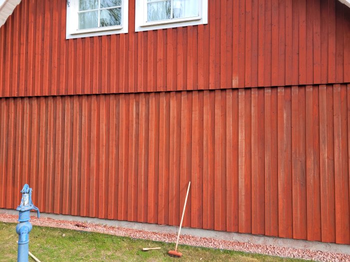 Rödfärgad träfasad på ett hus med vita fönster, en blå vattenpost och en soplucka nära marken.