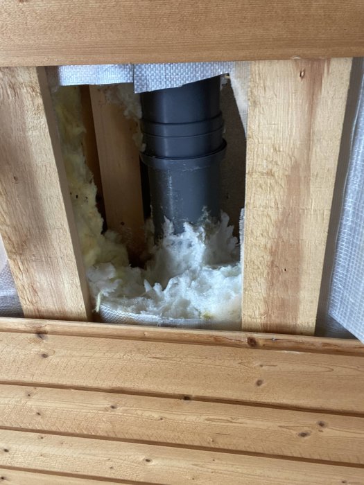 Avloppsrör från toalett med omgivande skumisolering synligt under trägolv.