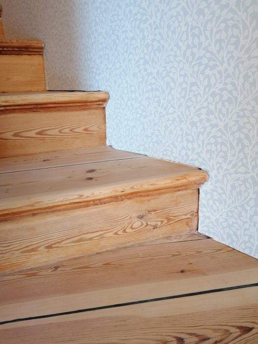 Detaljbild av trätrapp och golvsockel mot mönstrad tapet.