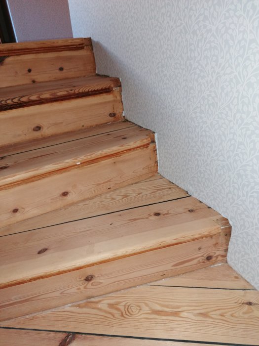 Trappa med obehandlade trästeg vid en tapetserad vinkelvägg, utan golvsockel.