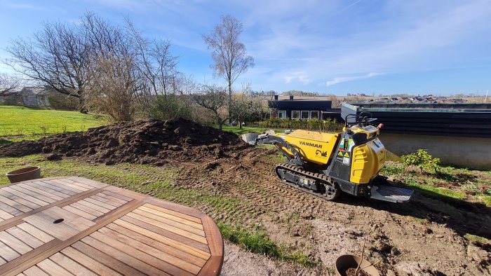 En gul Yanmar minidumper vid en hög med jord i en trädgård, bredvid gräsmattan och trädäck.