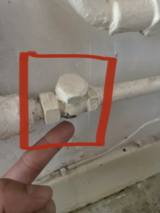 En hand pekar på en gammal vattenkoppling markerad med en röd ram på en vägg, vilket antyder byte eller underhåll.