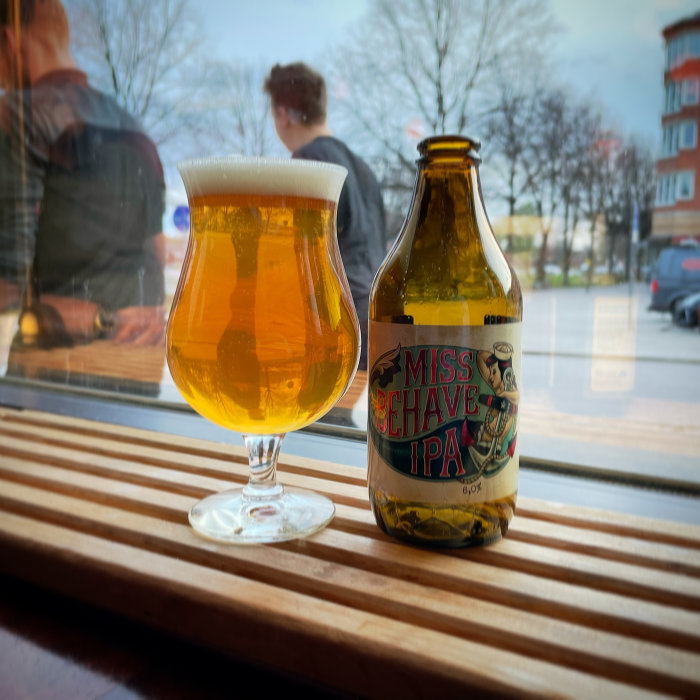 IPA öl i glas bredvid en ölflaska på ett ribbat bord med fönsterutsikt mot gatan.