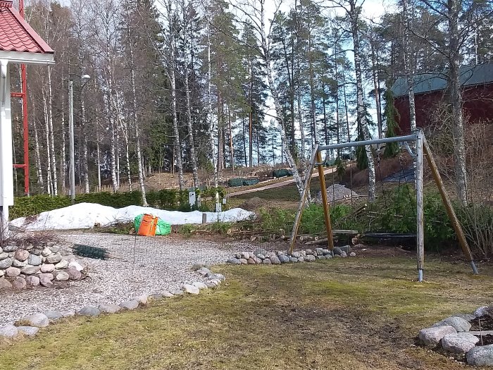 Trädgård med nyligen rensat område längs thuja-häck, gräsmatta i förgrunden, gungställning och skog i bakgrunden.