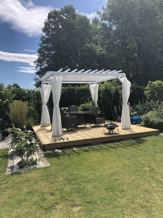 Nytt trädäck med pergola, sittgrupp och vita draperier i soligt trädgårdsmiljö.