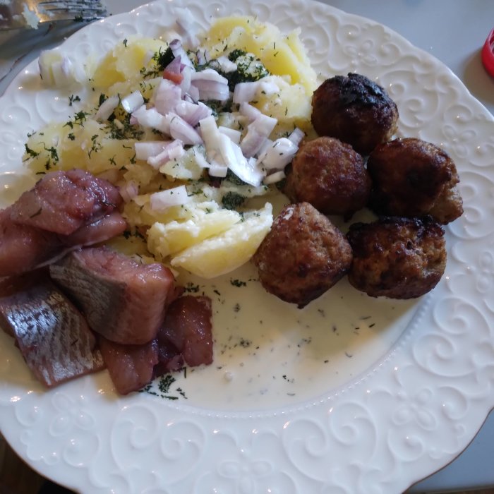 Traditionellt svenskt mål med sill, potatis, hackad lök och köttbullar på en vit tallrik.