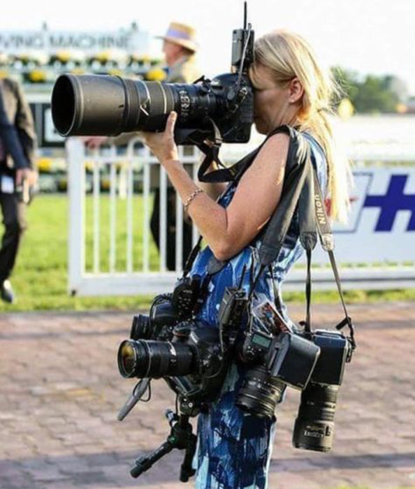 Kvinna på hästkapplöpningsbana fotar med stort objektiv och bär flera kameror.