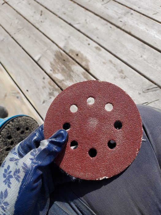 Hand med skyddshandske håller ett sliprondell med slitet kardborreludd och ett fäst sandpapper ovanpå en trätrall.