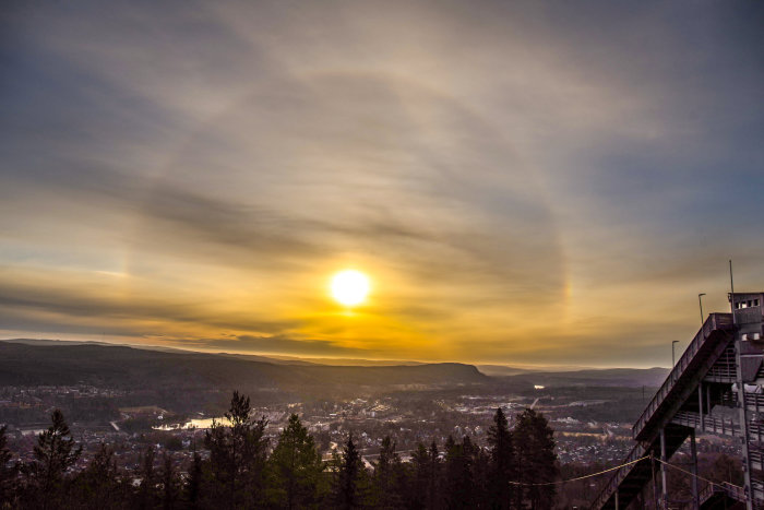 Halo-fenomen runt solnedgången över en stad med utsikt från ett skidhoppstorn, med berg och skog i bakgrunden.