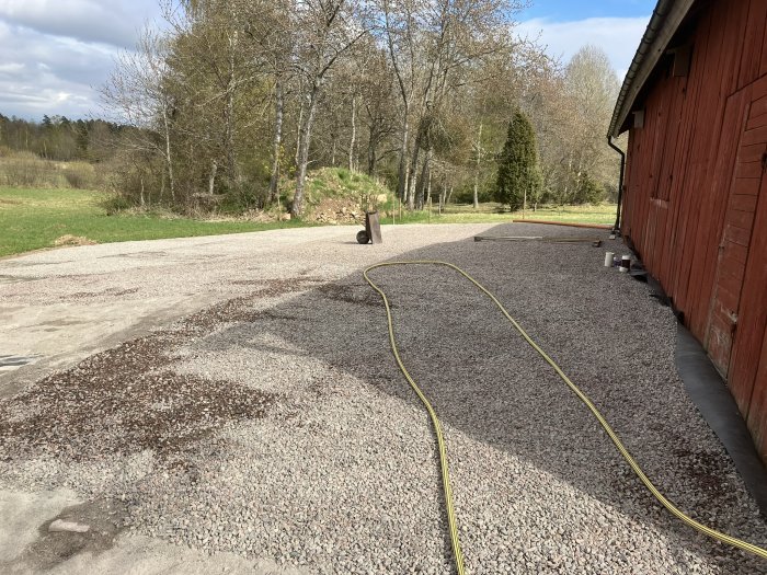 Gruslagd uppfart vid röd lada ett år efter anläggningen med gröna träd i bakgrunden och en vattenslang på marken.