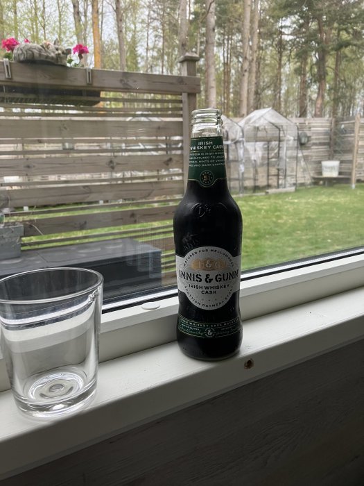 Flaska av Innis & Gunn öl på fönsterbräda med ett tomt glas och utsikt över trädgård.