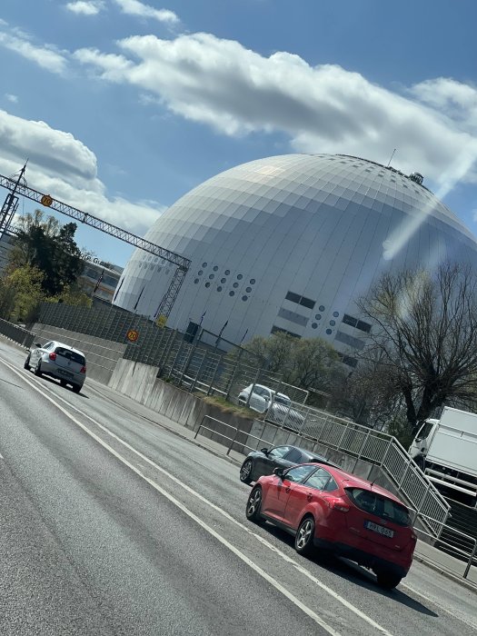 Kupolformad byggnad vid sidan av en väg med bilar i förgrunden och delvis molnig himmel ovanför.