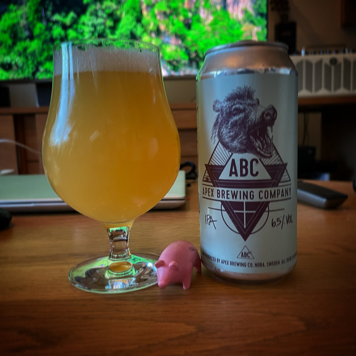 Ölglas med grumlig öl bredvid en ölburk från Apex Brewing och en liten rosa plastgris framför en skärm.