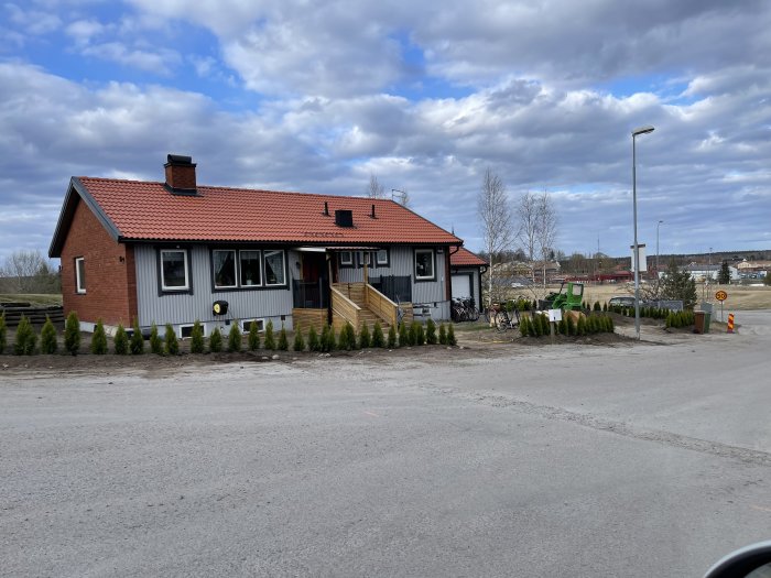 Nyligen planterade thujor framför en tegelröd villa med molnig himmel i bakgrunden.