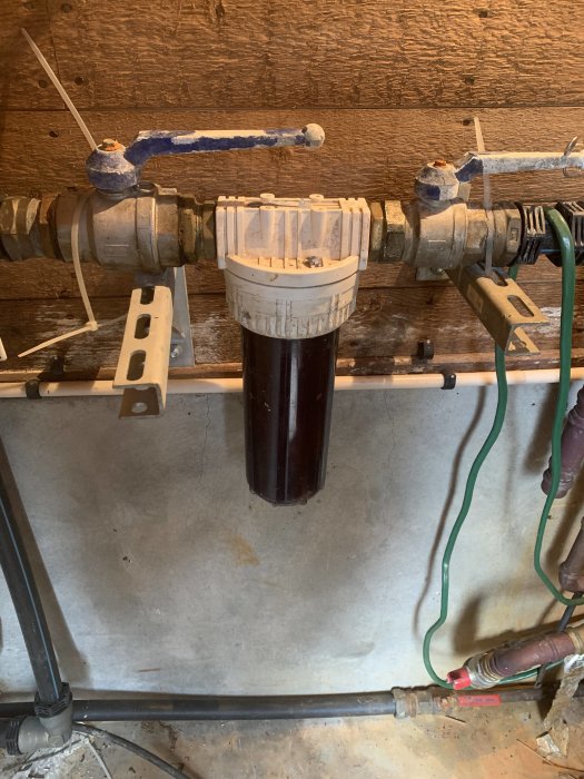 En pumpmontering med rör, en vit topp och en mörk cylinder på en trävägg i ett installationsrum.