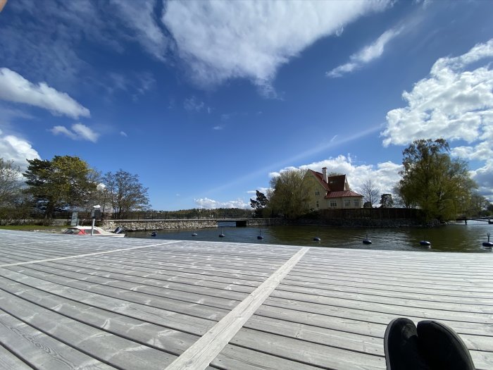 Träbrygga behandlad med Organowood vid vattnet, med hus och träd i bakgrunden under en klar himmel.