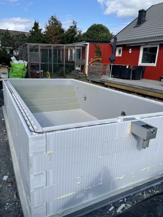 En nyligen installerad vit pool i en trädgård med röd husfasad och växthus i bakgrunden.