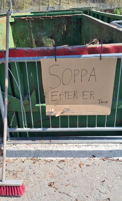 En wellpappskylt med texten "SOPPA EFTER ER Tack" fäst vid en grön container bredvid en röd sopkvast.