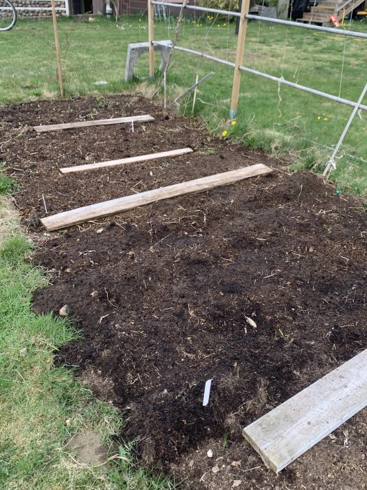 Planteringbäddar med jord förberedda för växter, omgivna av gräs med staket i bakgrunden.