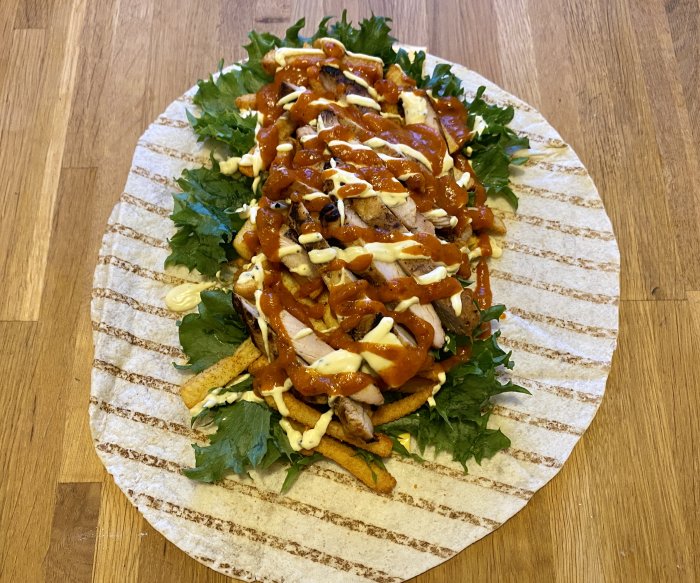 Överfylld wrap med ost, sallat, pommes, kyckling och såser på ett köksbord.